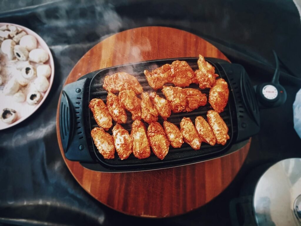Top view of delicious spicy chicken wings grilling on an electric grill with mushrooms nearby.
